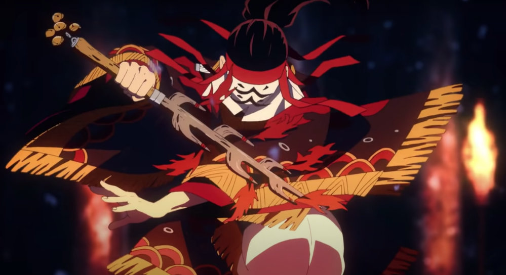 Screengrab of a heavily costumed figure in the middle of a dance. The figure's face is covered in a white veil with black marks, and is wearing costume brightly coloured in reds and vivid orange. The figure is holding a wooden pole with pointed ends on one side and brass bells on the other.