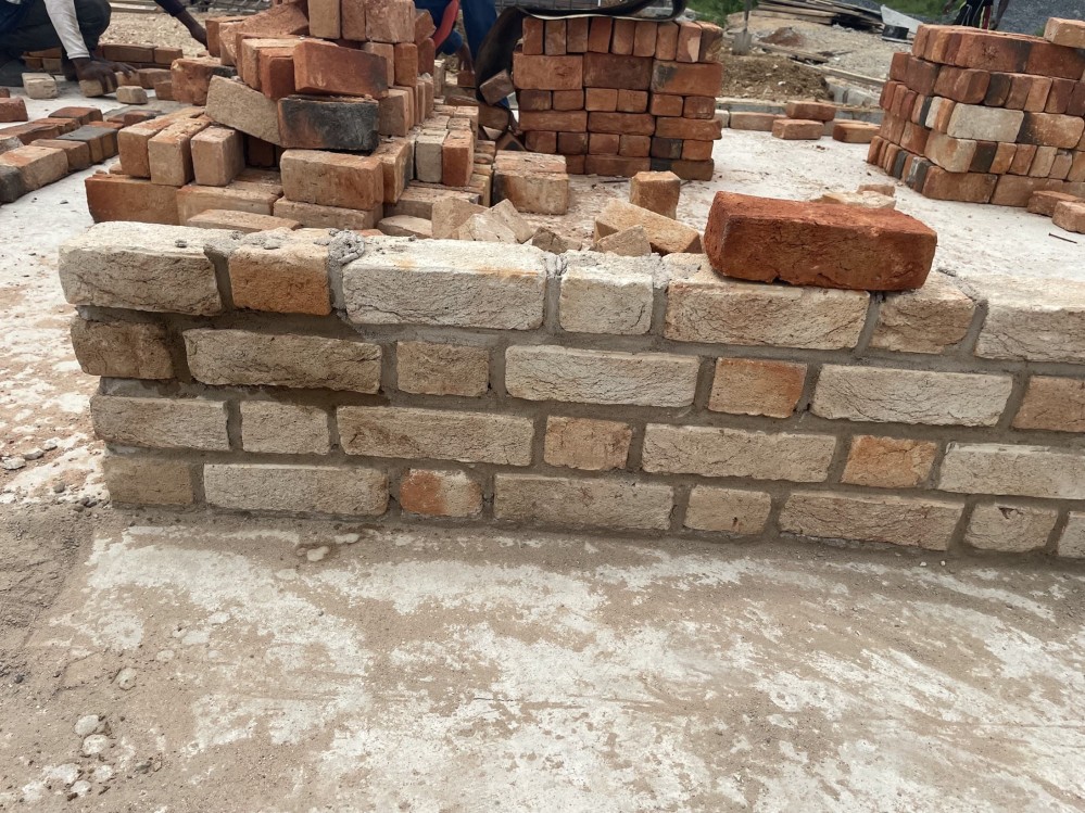 A small sample wall, four bricks high, made of light brown and tan bricks. A single deep red brick sits on top.