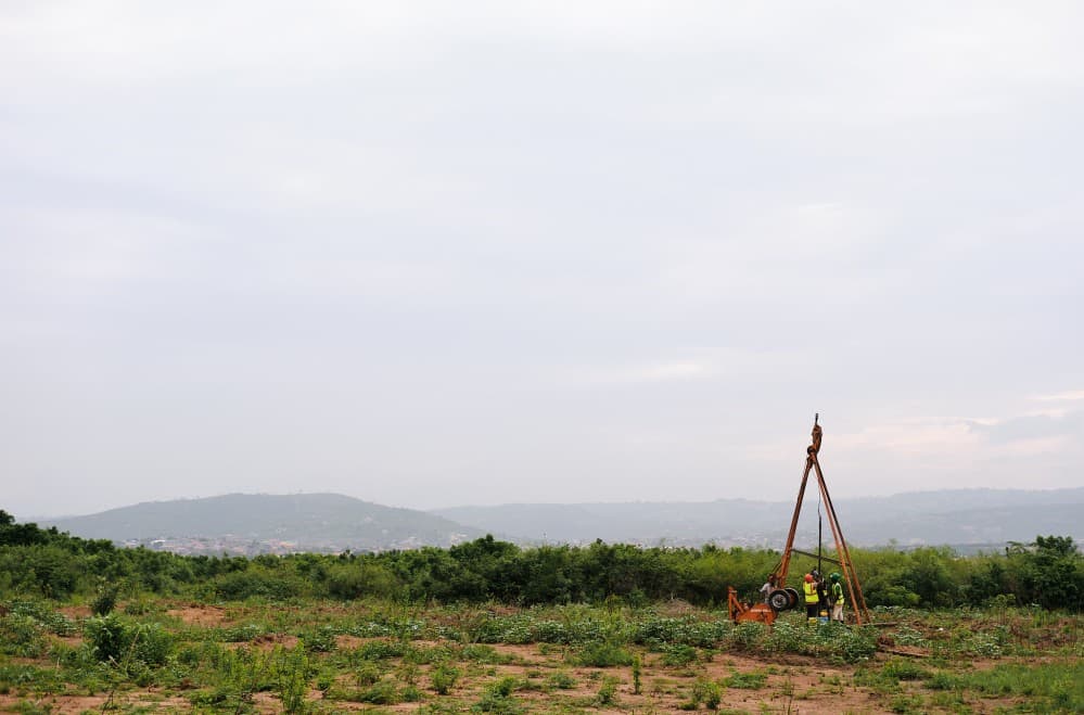Wide shot of three workers drilling into the ground. They're small figures at the bottom right of the photo, almost swallowed by the large surrounding landscape