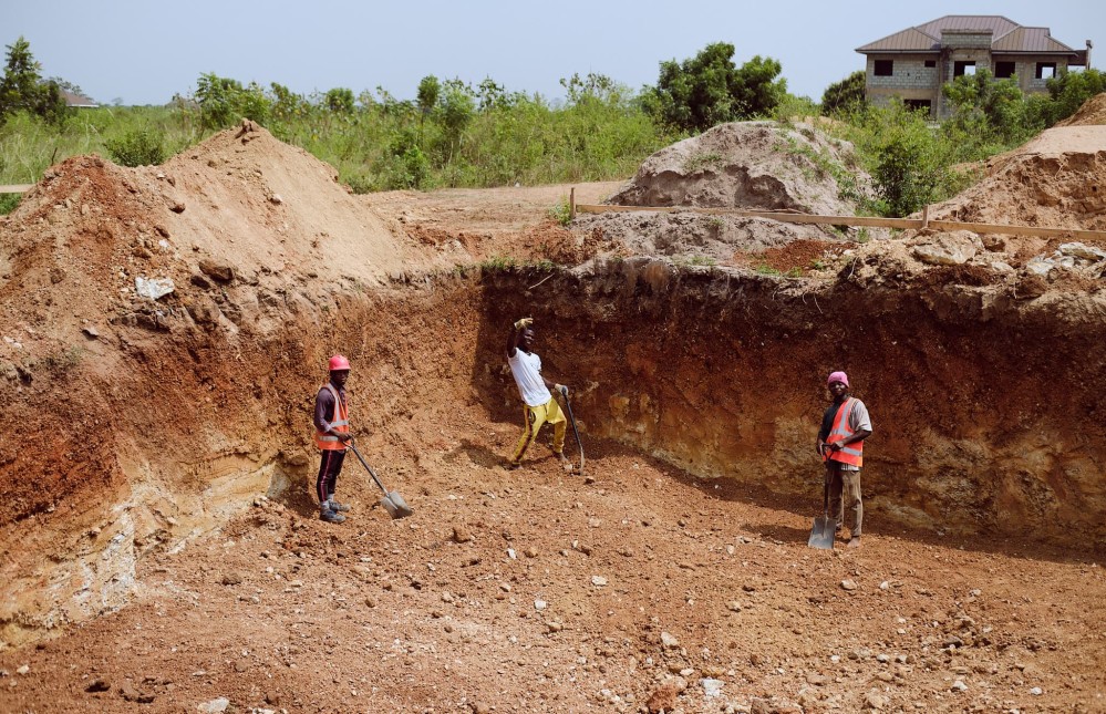 Three workers wave from the bottom of the excavation. The excavation is about three heads taller than an average height man.