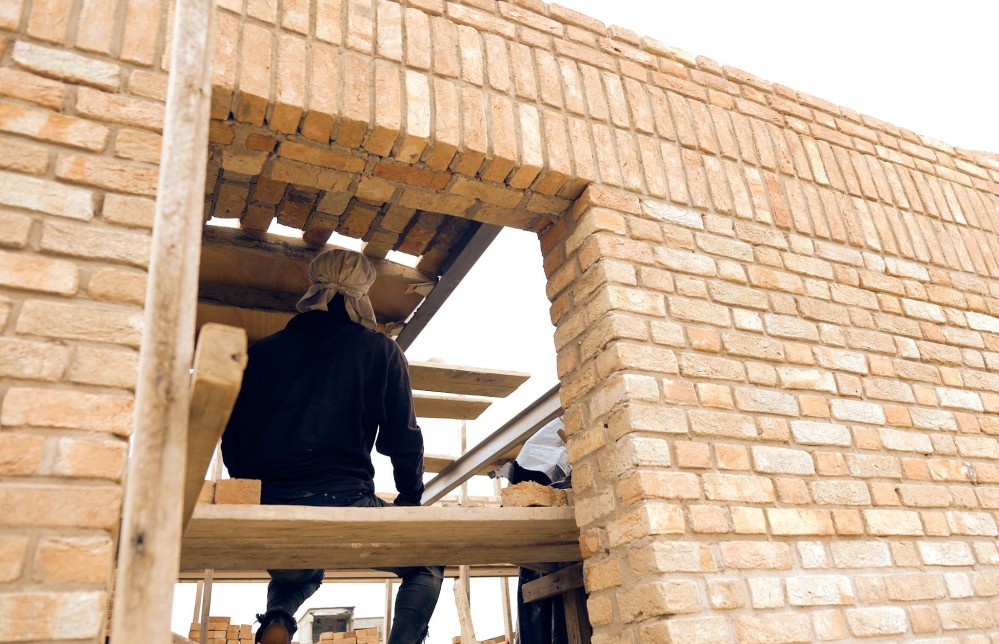 A worker sits on a plank above ground, completing the barell-vaulted ceiling. You can see him through a brick doorway