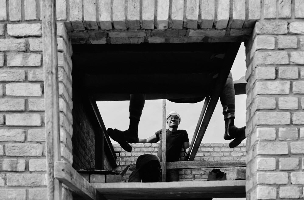 Black and white photo of a view through brick doorway, showing the dangling legs of a bricklayer in the foreground, or a smiling colleague in the background