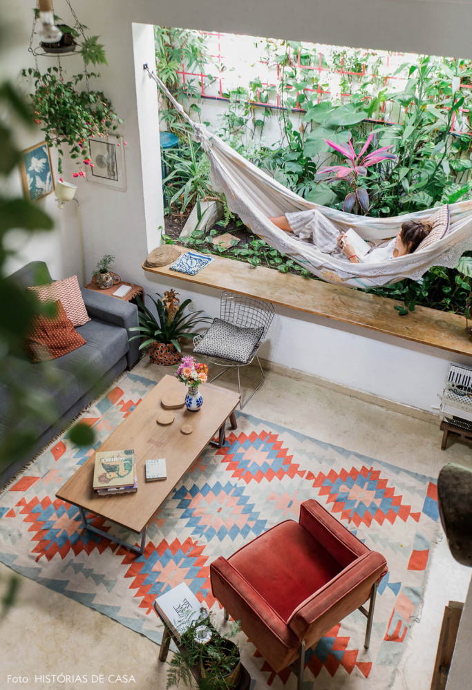 A living room with an unusual feature - there is large window area without glass. Instead, a hammock is slung in the space, which opens into a leafy side yard. The living room itself seems modest/ There's a small dark grey couch, a small rectangular wooden table, and a large rug patterns in geometric reds, blues, and pinks. A small red armchair is in the bottom right of the image.