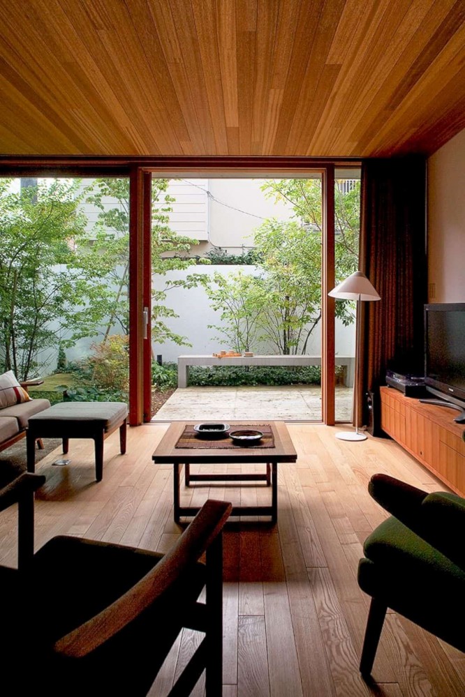 Dark room with wooden floor and wooden ceiling, opens into a very bright sideyard with green shrubs and a small concrete bench.