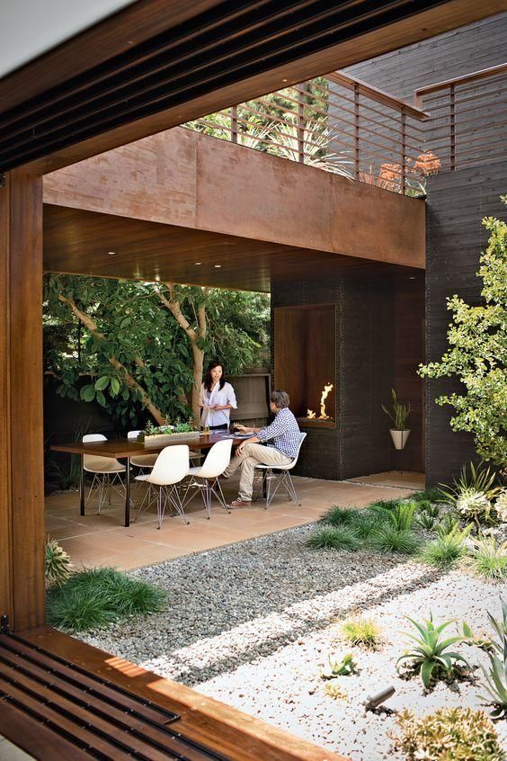 Large dining area that hugs an open courtyard. A lady and gentleman appear in conversation at the table. The courtyard has grey gravel and several shrubs and tall tree. Another interesting element is what appears to be a rusty steel terrace above the eating area, looking into the courtyard