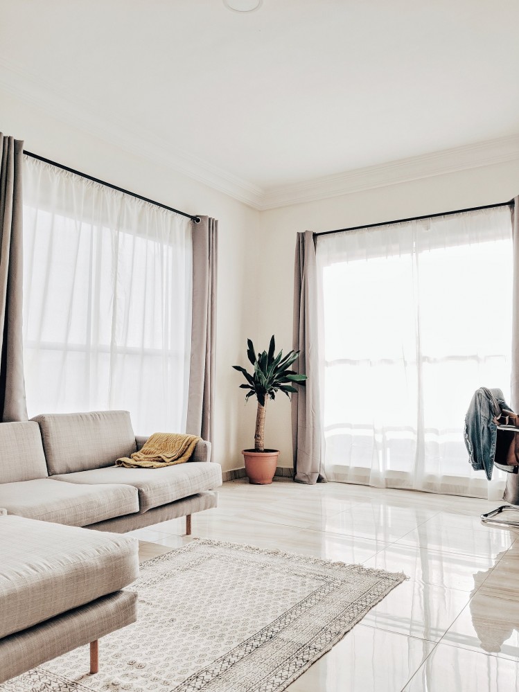 Bright white living room in neutral tones with sheer curtain windows running to the bottom. A grey couch rests on a grey carpet on a grey tile floor. A yellow blanket on the couch and short tree in a terra cotta pot in the corner provide some colour.