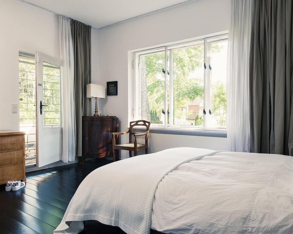 Small bed with white bedsheets resting on what looks like black lacquered wood panels. Light is streaming through window on the right hand side and through a door facing the bed. There are brown wood accents from two dressers and a bedside chair, and