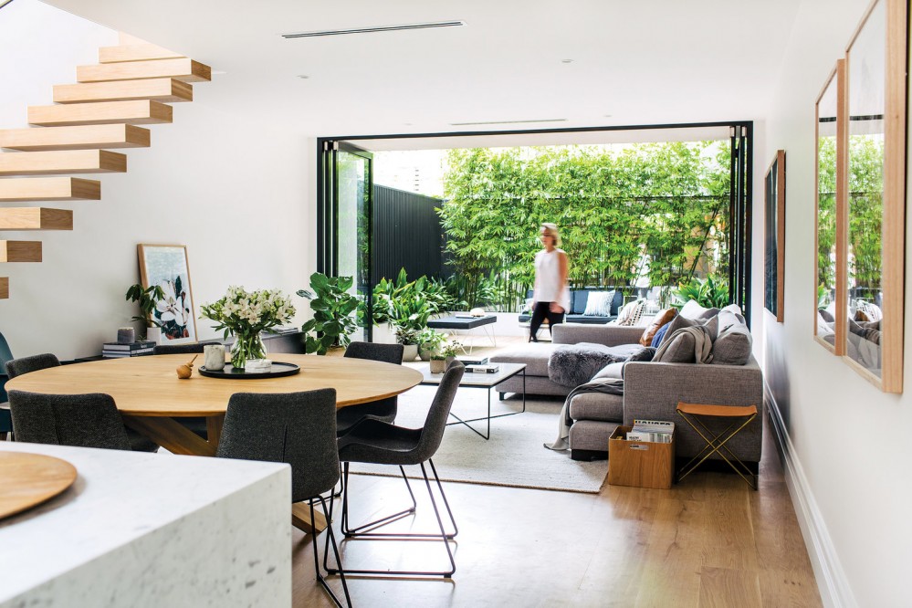Photo of a large open concept space that appears to be a combination dining and living area. It opens up into an enclosed outdoor area. One wall appears to be black painted wood, and the other wall is covered with greenery, maybe bamboo shoots.